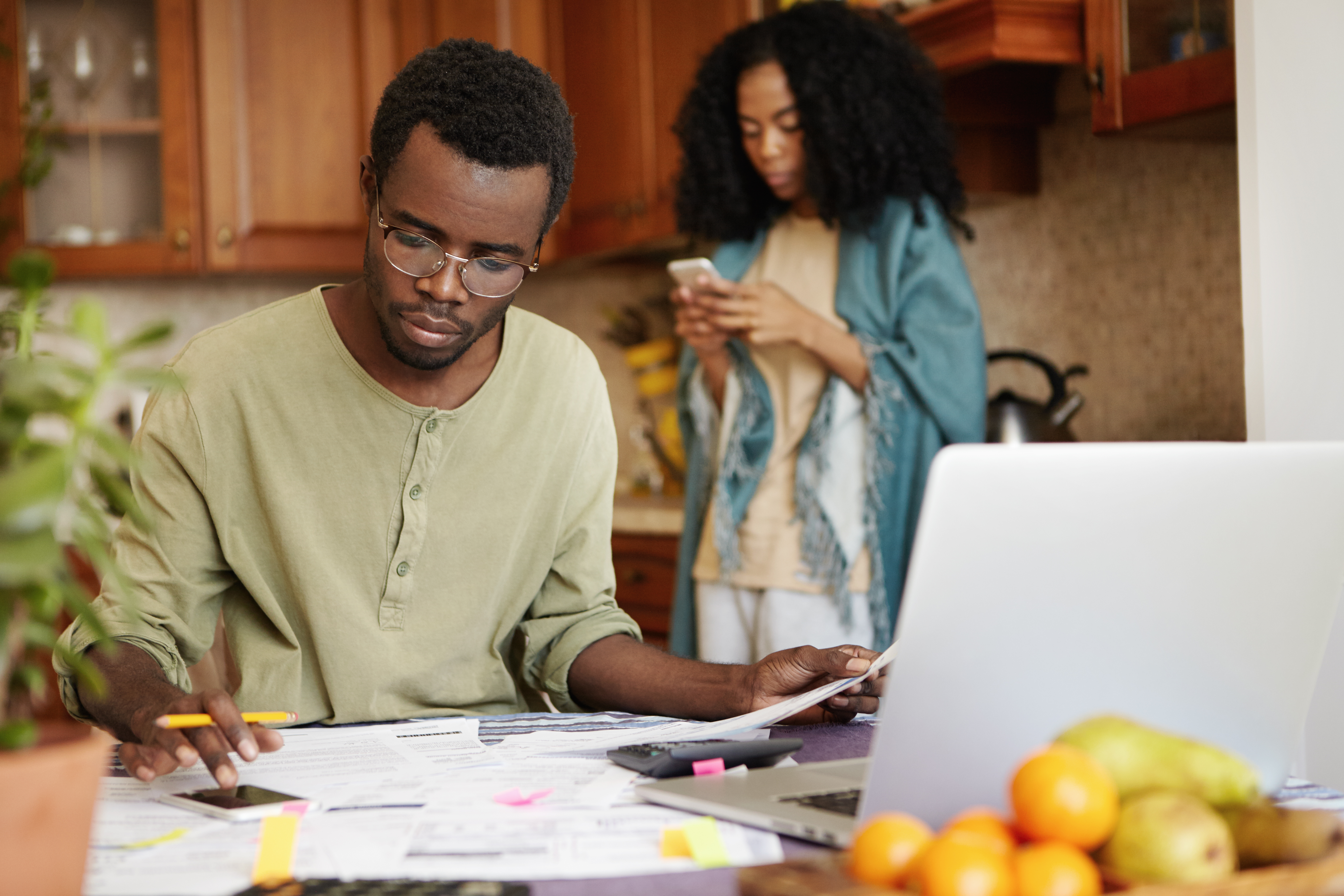 man sitting in kitchen looking at paper using a calculator with a woman on her mobile phone
