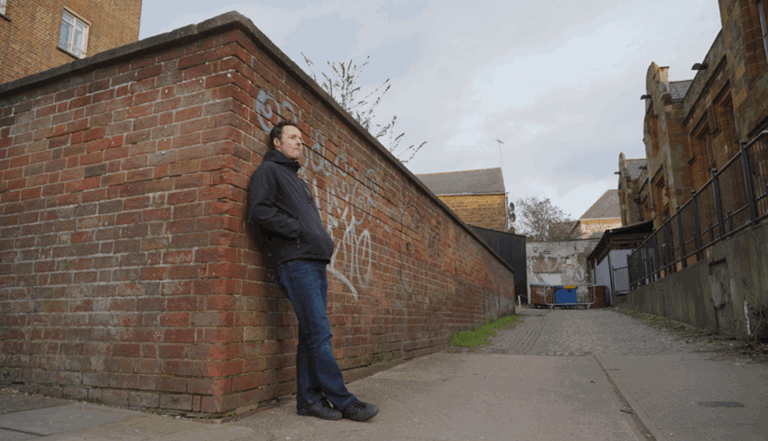Full body view of a man named Kieran leaning against a wall with graffiti wearing a black jacket and blue jeans Full body view of a man named Kieran leaning against a wall with graffiti wearing a black jacket and blue jeans